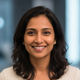 Headshot portrait of a happy South Asian woman smiling confidently, with a soft slate blue and white background.